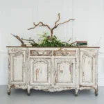 A vintage, weathered wooden sideboard adorned with greenery and decorative branches sits against a bright, white wall.