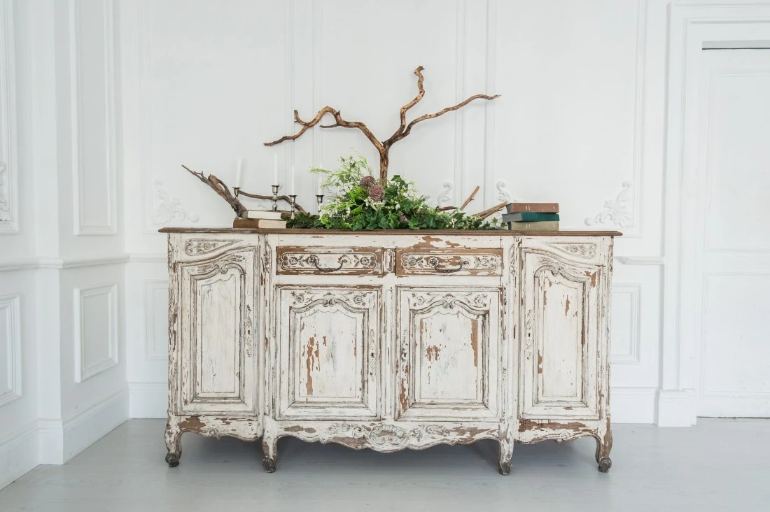 A vintage, weathered wooden sideboard adorned with greenery and decorative branches sits against a bright, white wall.