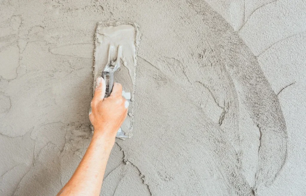 A hand holds a trowel, smoothing wet concrete on a textured wall surface, showcasing the process of wall finishing.