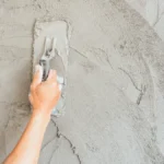 A hand holds a trowel, smoothing wet concrete on a textured wall surface, showcasing the process of wall finishing.