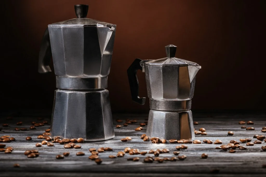 A large and small metal stovetop espresso maker sit on a wooden table scattered with coffee beans, against a dark backdrop.