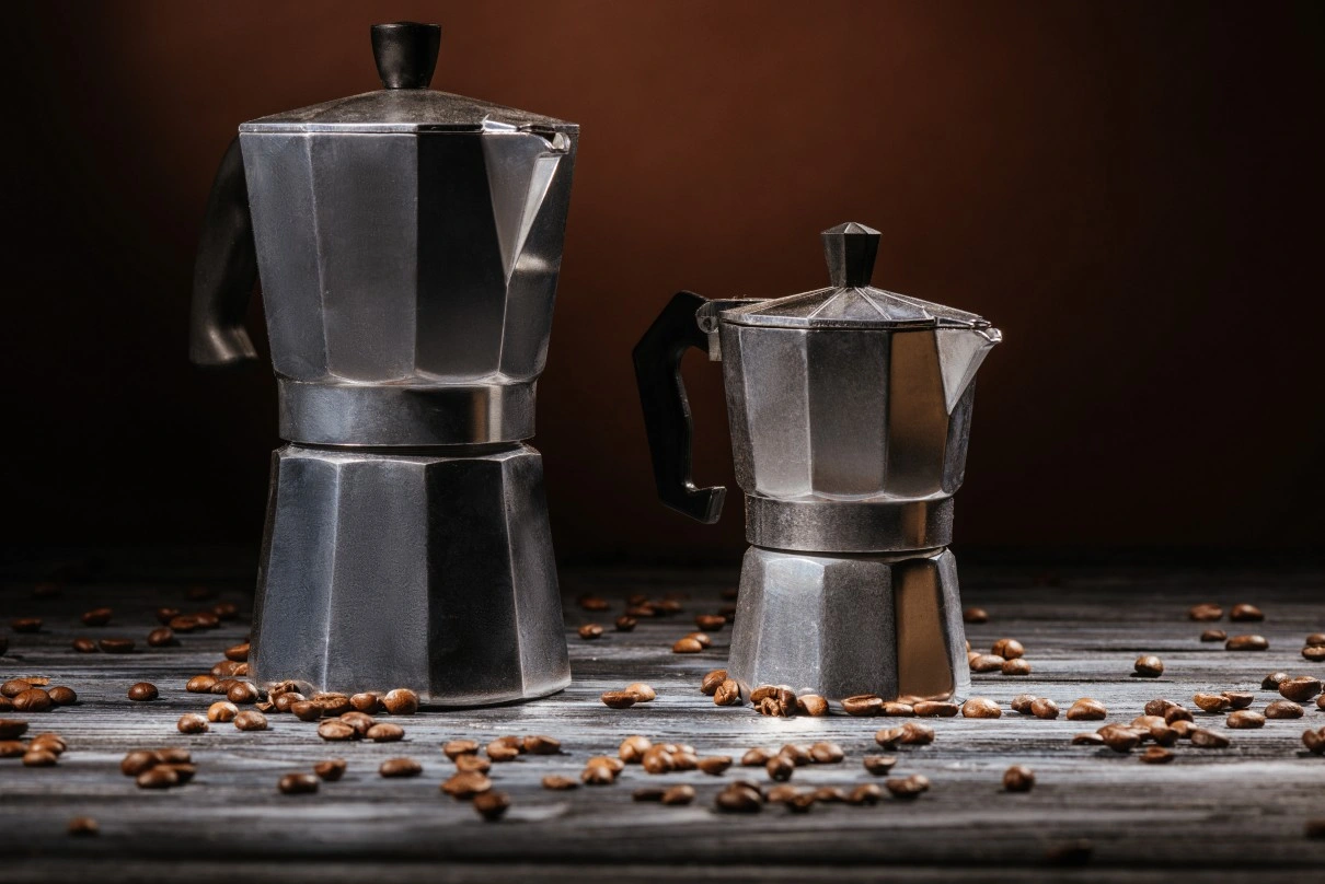 A large and small metal stovetop espresso maker sit on a wooden table scattered with coffee beans, against a dark backdrop.