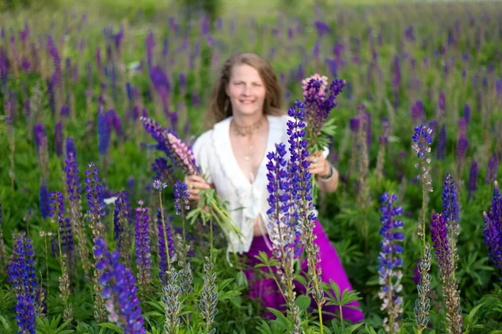 A woman smiles joyfully amidst a vibrant field of purple lupines, holding several colorful flower stems in her hands.