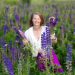 A woman smiles joyfully amidst a vibrant field of purple lupines, holding several colorful flower stems in her hands.