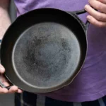 A person holds a well-used cast iron skillet, showcasing its dark, seasoned surface, wearing a purple shirt and plaid shorts.