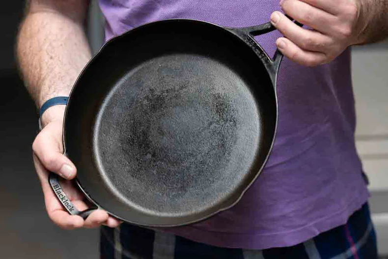 A person holds a well-used cast iron skillet, showcasing its dark, seasoned surface, wearing a purple shirt and plaid shorts.