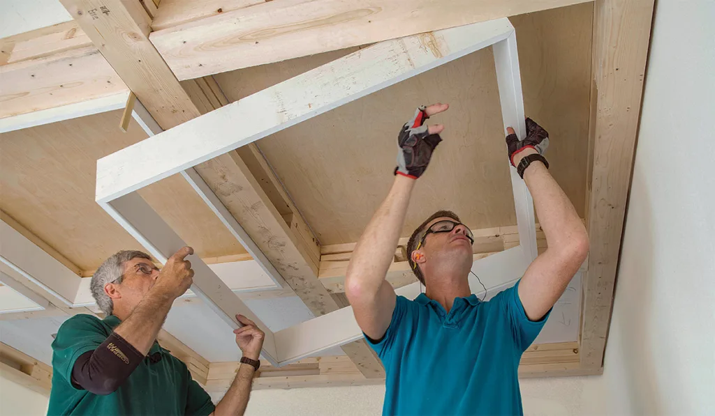 Two men work together, installing a white frame onto a wooden ceiling structure, focused and using tools for precision.