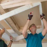Two men work together, installing a white frame onto a wooden ceiling structure, focused and using tools for precision.