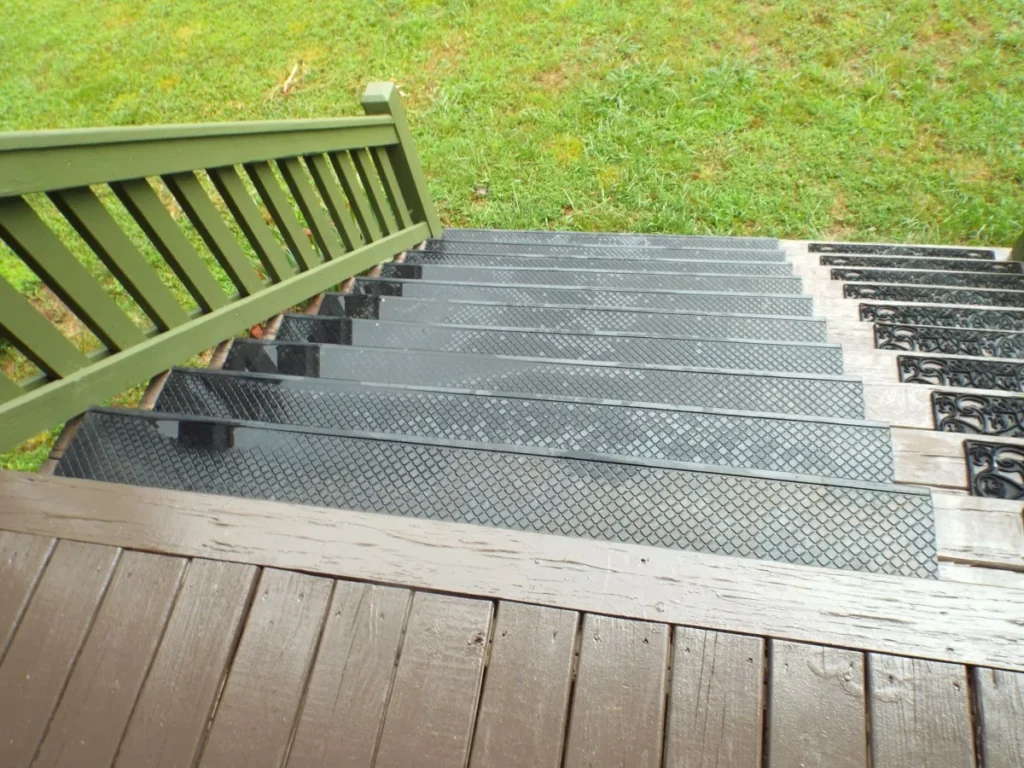 View looking down a set of black rubber stairs treads with diamond-patterned treads and a green wooden railing, leading to a grassy yard.