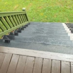 View looking down a set of black rubber stairs treads with diamond-patterned treads and a green wooden railing, leading to a grassy yard.