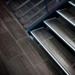 Close-up view of modern indoor stairs with dark stair treads and a metal railing, shot at an angle showing the steps and part of the adjacent tiled floor.