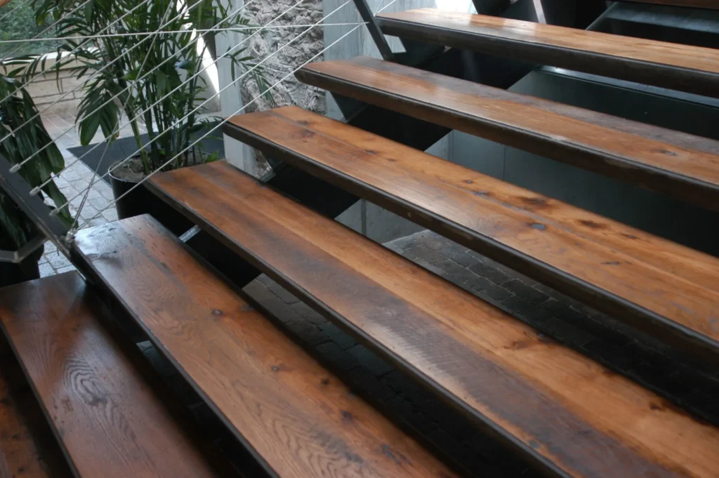 Close-up of polished wooden stair treads with metal supports and a railing featuring thin metal cables, next to potted green plants.