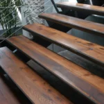 Close-up of polished wooden stair treads with metal supports and a railing featuring thin metal cables, next to potted green plants.