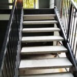 Concrete outdoor stairs with black metal railings on both sides, leading up to a landing with greenery and parked cars visible.
