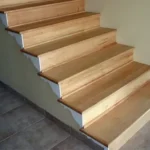 A wooden staircase with light-colored steps ascends beside a plain beige wall, adjacent to a tiled floor.
