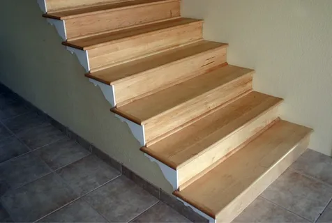 A wooden staircase with light-colored steps ascends beside a plain beige wall, adjacent to a tiled floor.