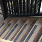 Wooden outdoor stairs with black anti-slip strips, bordered by black vertical railing, descending beside a deck.