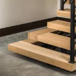 Close-up of modern floating wooden stairs with black metal railings against a concrete floor and white wall background.